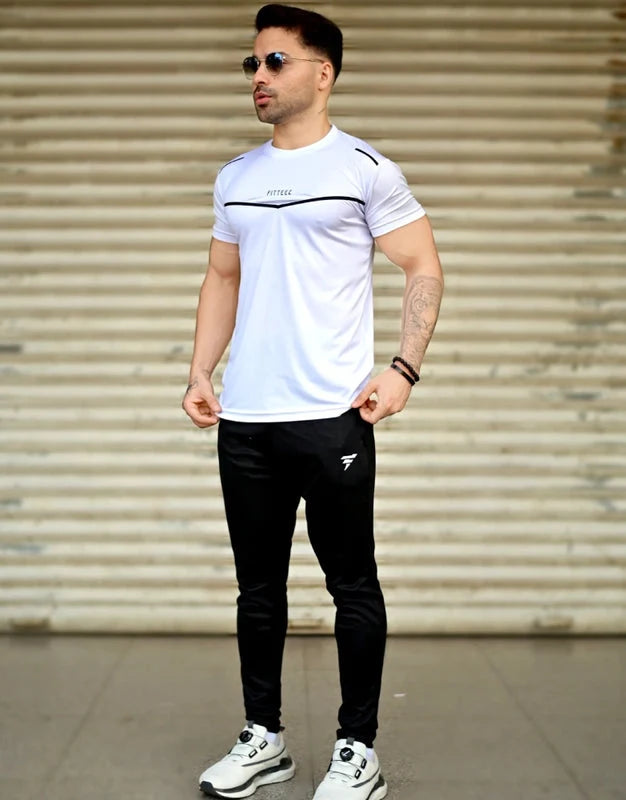 Man wearing a white t-shirt and black pants with visible branding, standing against a metallic shutter. Leisure Icon White & Black Track Suit Royal Outfits Track Suits