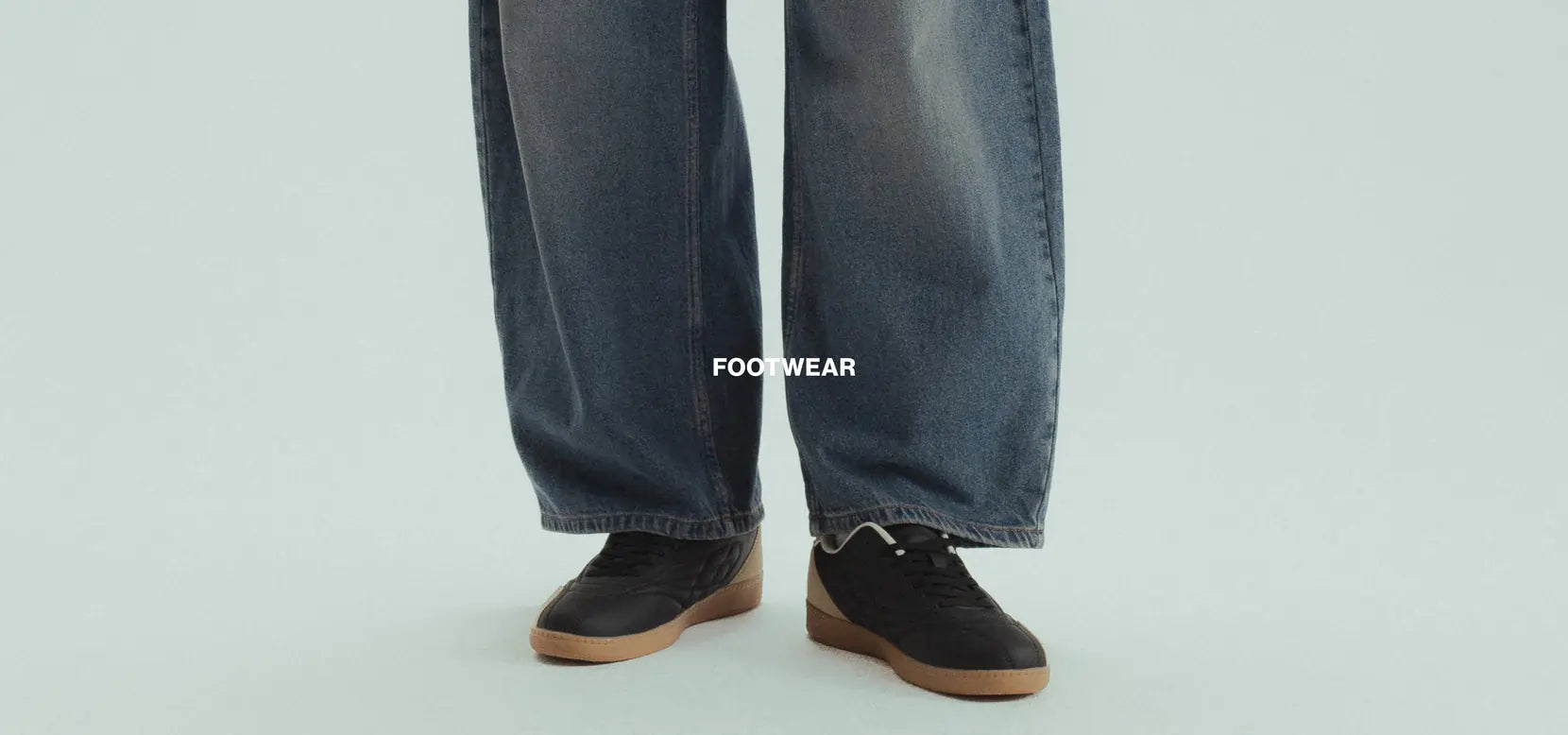 Person wearing dark jeans and black shoes with brown soles on a light gray background