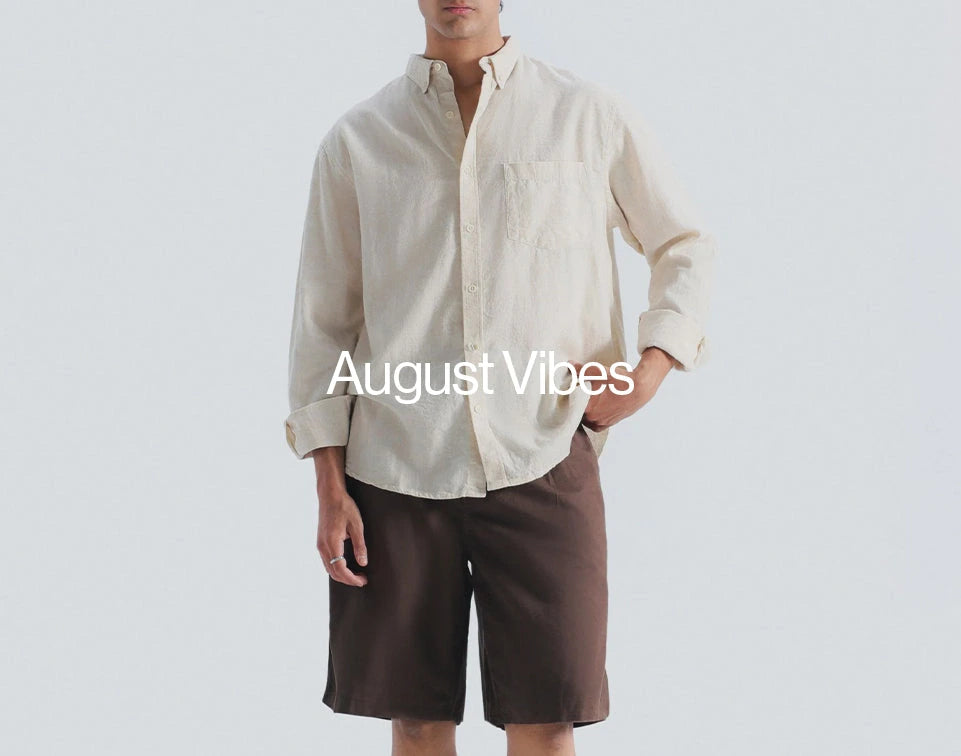 A man wearing a light beige long-sleeve button-up shirt and dark brown shorts, standing against a light gray background with the text 'August Vibes' overlaid across the center.