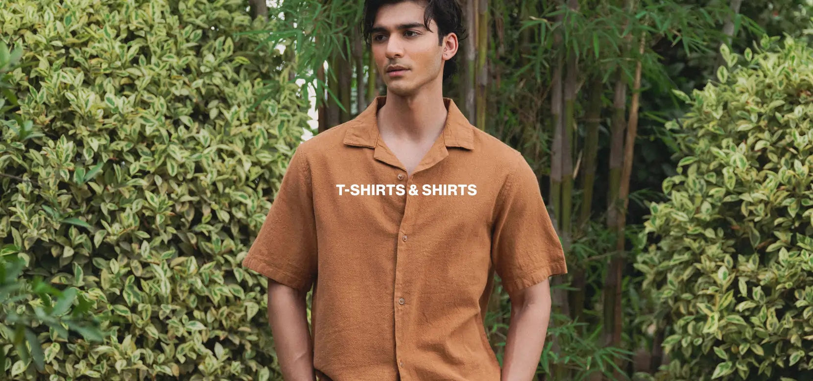 Man wearing a brown shirt with 'T-Shirts & Shirts' text standing in front of green foliage.